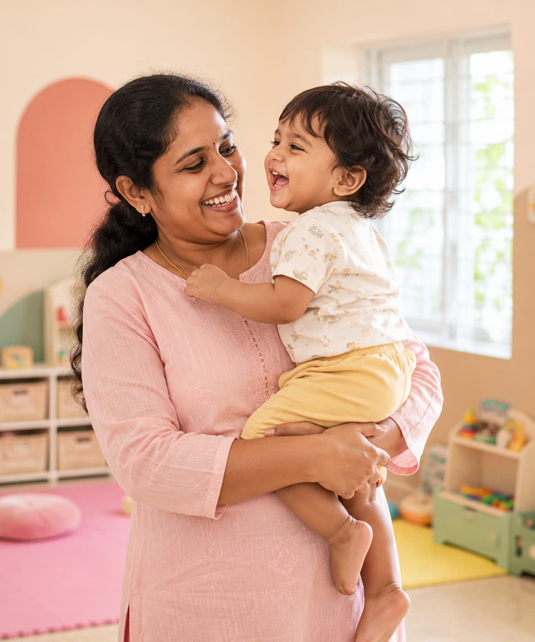 Caregiver smiling with a toddler on her hip in a bright, welcoming room at Tiny Cradle Puducherry
